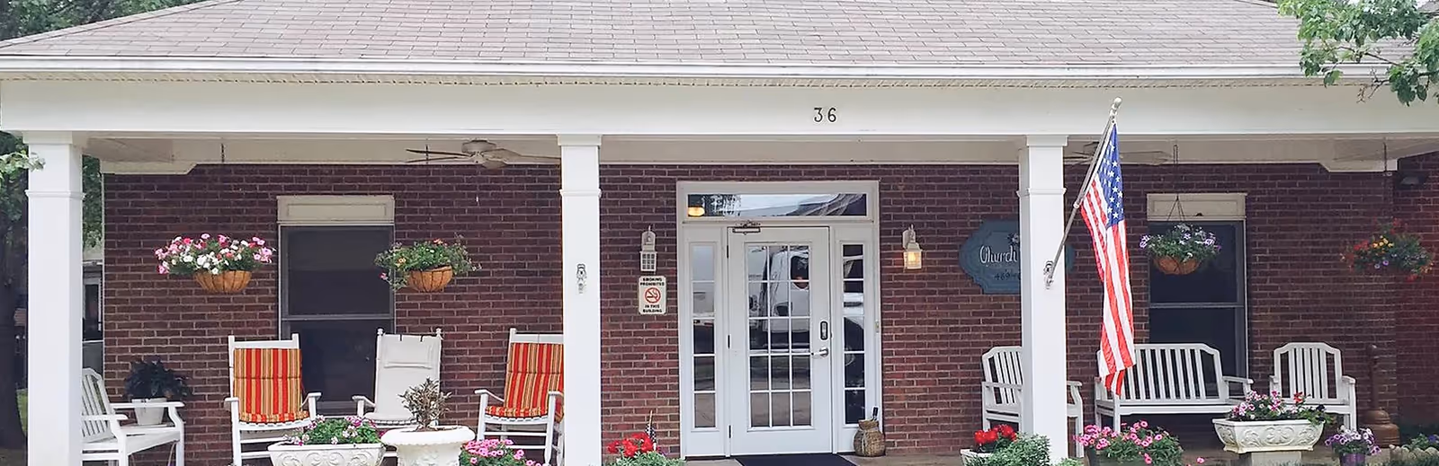 Brick building front with white columns, porch seating, hanging plants, and an American flag.