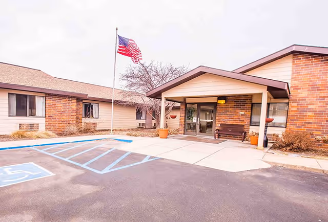 Exterior view of Centennial Park Retirement Village showing the entrance with a covered porch, a bench, potted plants, an American flag on a flagpole, and a parking area with a handicapped parking space.