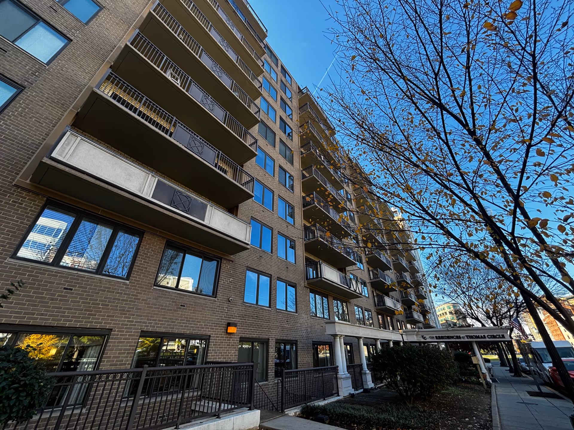 Low-angle view of a multi-story brick apartment building with balconies, an entrance canopy, sidewalk, and a tree against a blue sky.