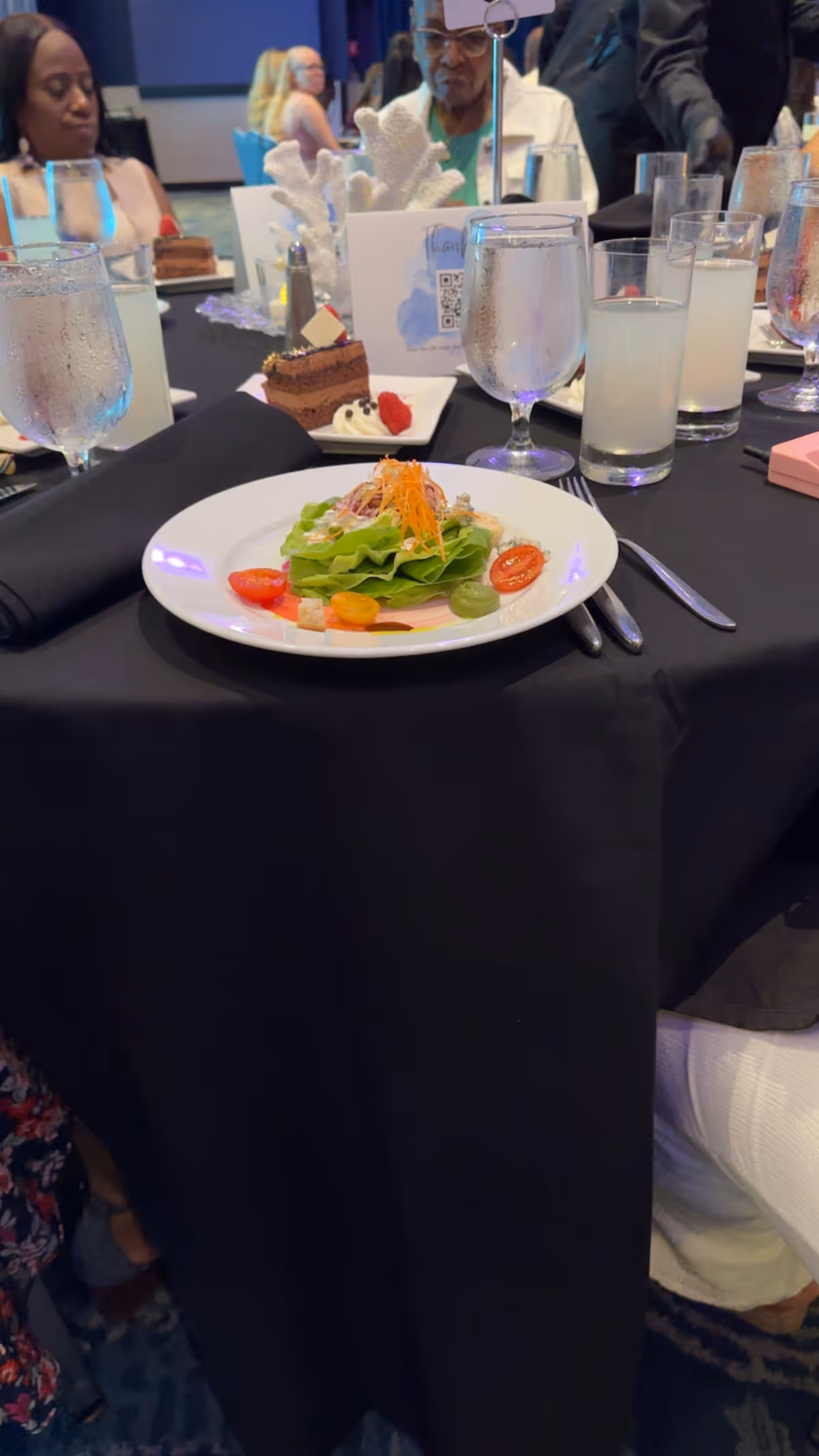 Table set with salad, desserts, and drinks; guests seated at a formal event.