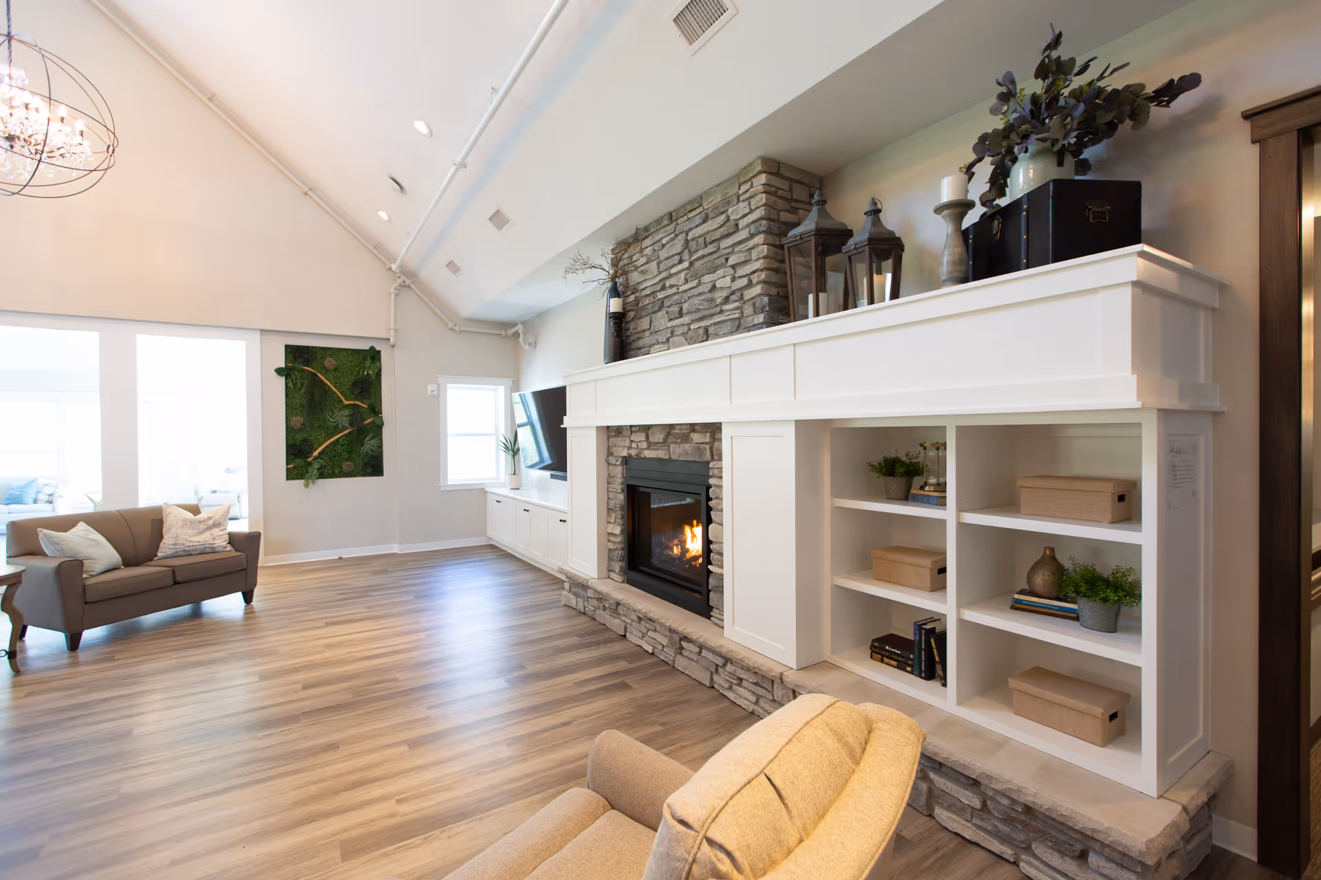 A spacious living room with light wood flooring, a stone fireplace with a white mantel, built-in shelves with decorative items, a mounted flat-screen TV, a gray loveseat with pillows, and a beige armchair. The room has high ceilings with exposed pipes and a large window letting in natural light.