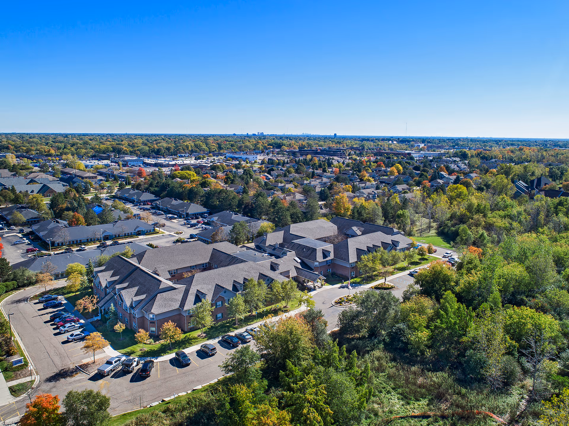 Aerial view of Comfort Cove Premier Senior Living facility surrounded by trees and greenery, with parking lots and nearby residential areas under a clear blue sky.
