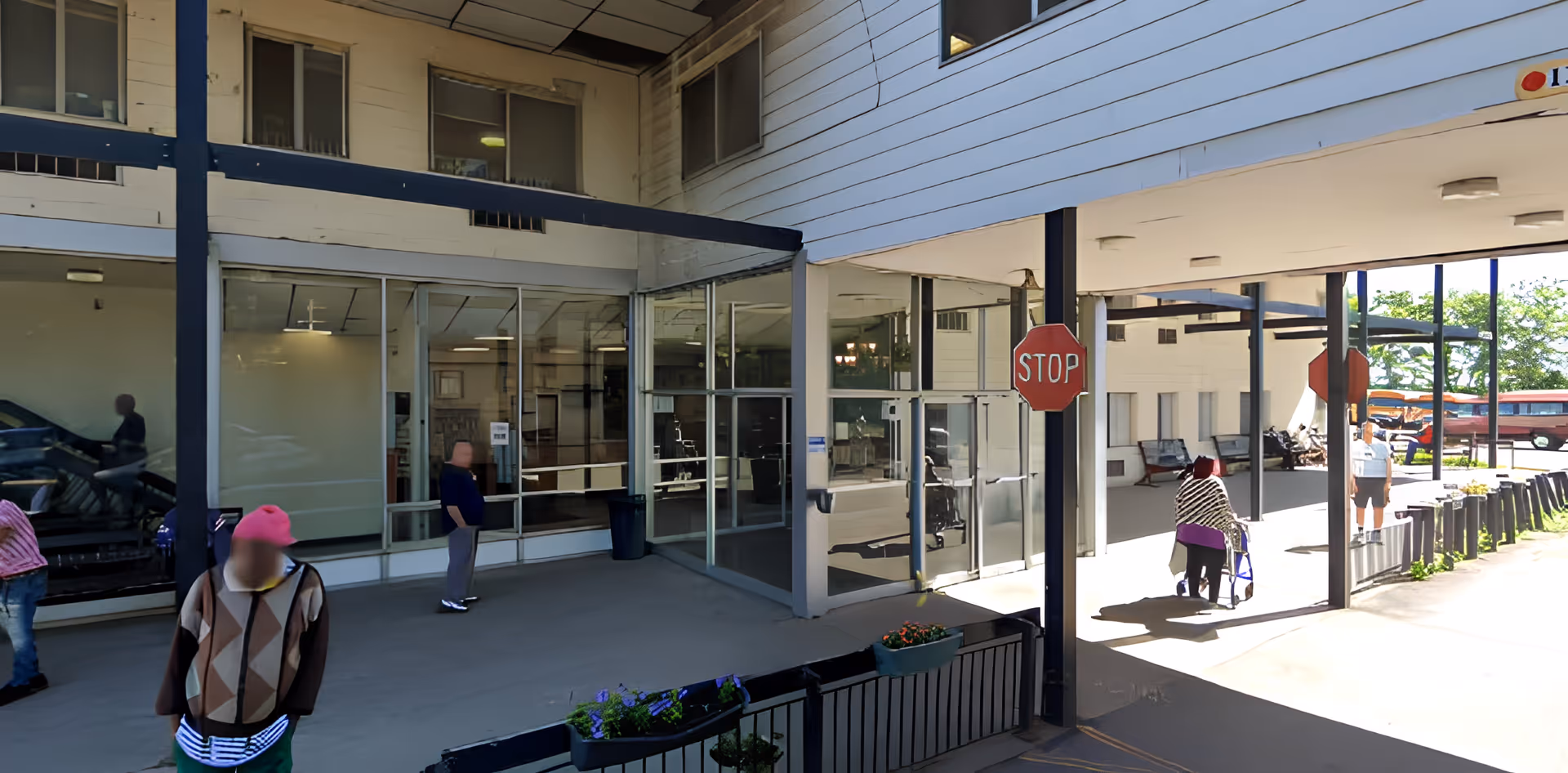 Covered glass entrance of a senior living facility with a stop sign and several people standing or using walkers nearby.