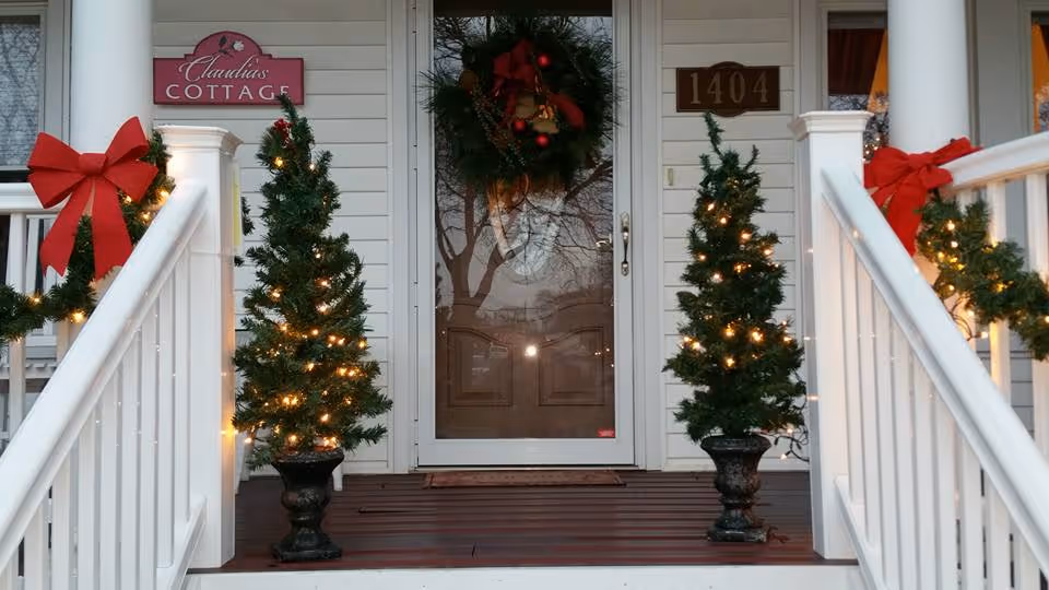 Front porch of Claudia's Cottage decorated for Christmas with two small lit evergreen trees, a wreath, red bows, and house number 1404.