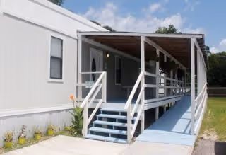 Exterior view of a single-story modular building with a covered porch and ramp entrance. The building is white with several windows, and there are small plants along the foundation. The sky is clear with some clouds.