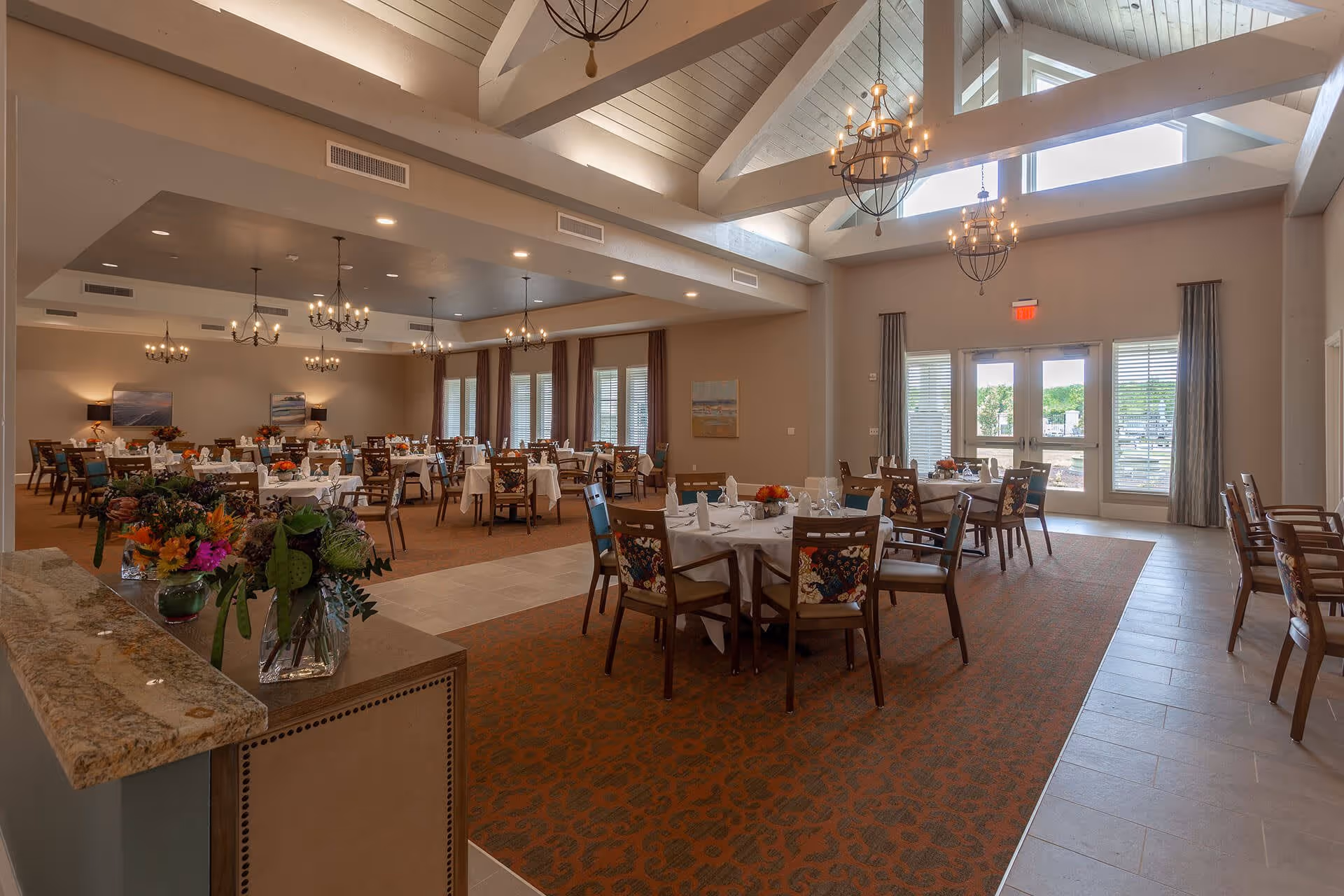 A spacious dining room with multiple round tables covered with white tablecloths and set with glassware and napkins. The room features high vaulted ceilings with exposed beams and multiple chandeliers. Large windows and glass doors allow natural light to fill the space. Floral arrangements are placed on a counter in the foreground.