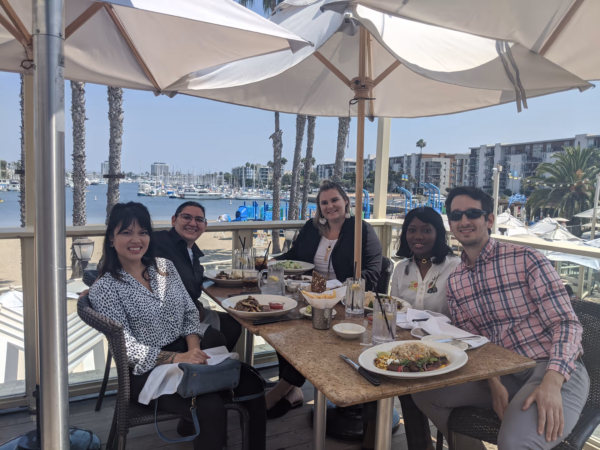 Five people dining outdoors at a marina restaurant with umbrellas.
