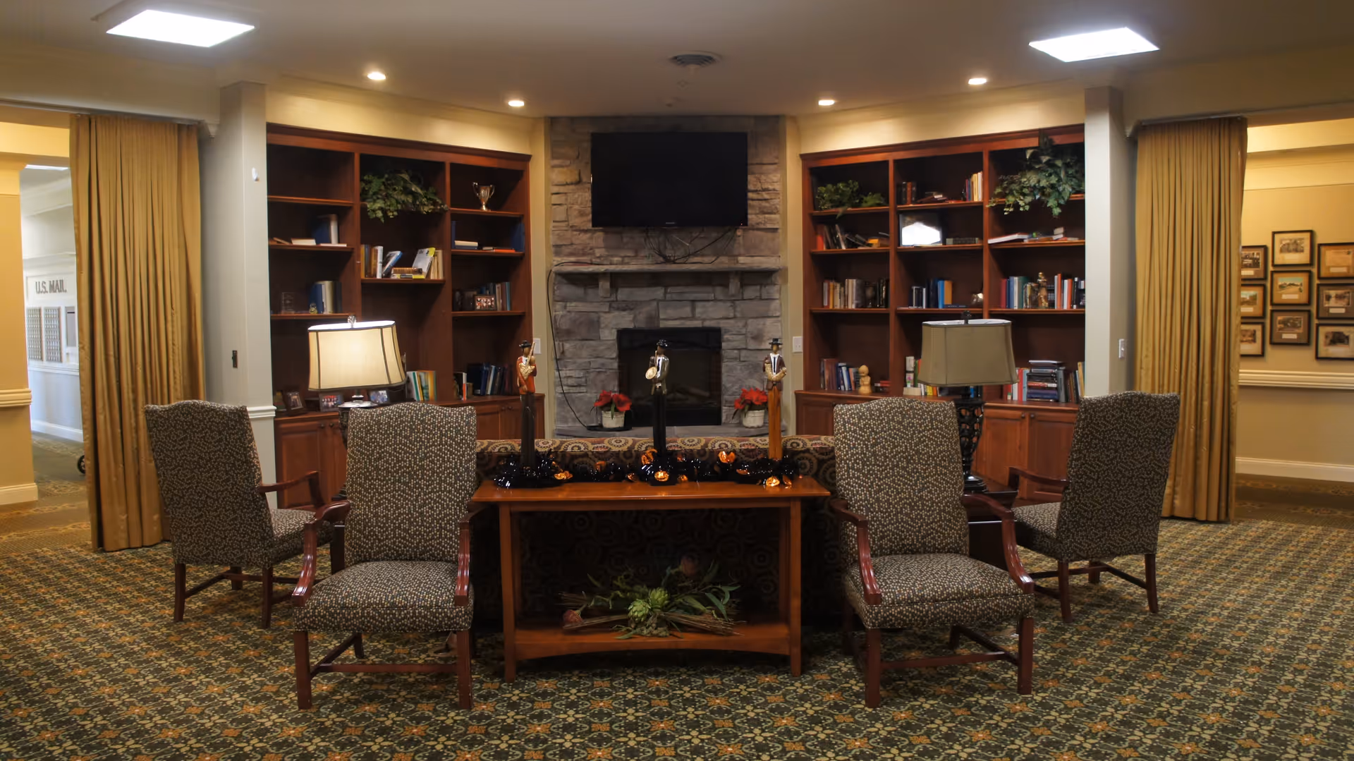A cozy common area with patterned armchairs arranged around a wooden table decorated with small figurines and Halloween-themed garland. Behind the table is a stone fireplace with a mounted flat-screen TV above it, flanked by built-in wooden bookshelves filled with books and plants. The room has warm lighting and carpeted floors with a patterned design.