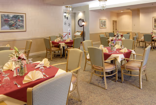 A dining room with multiple tables set for a meal. Each table has a red tablecloth with a white overlay, folded napkins, glassware, silverware, and floral centerpieces. The room has beige walls with framed artwork and carpeted flooring. Several chairs with light upholstery surround the tables.