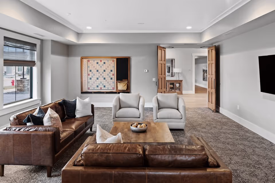 A cozy living room with two brown leather sofas and two light gray armchairs arranged around a wooden coffee table with a bowl of decorative balls. A large Scrabble board is mounted on the wall, and there is a window with blinds on the left side. Double wooden doors lead to another room, and a flat-screen TV is mounted on the right wall.