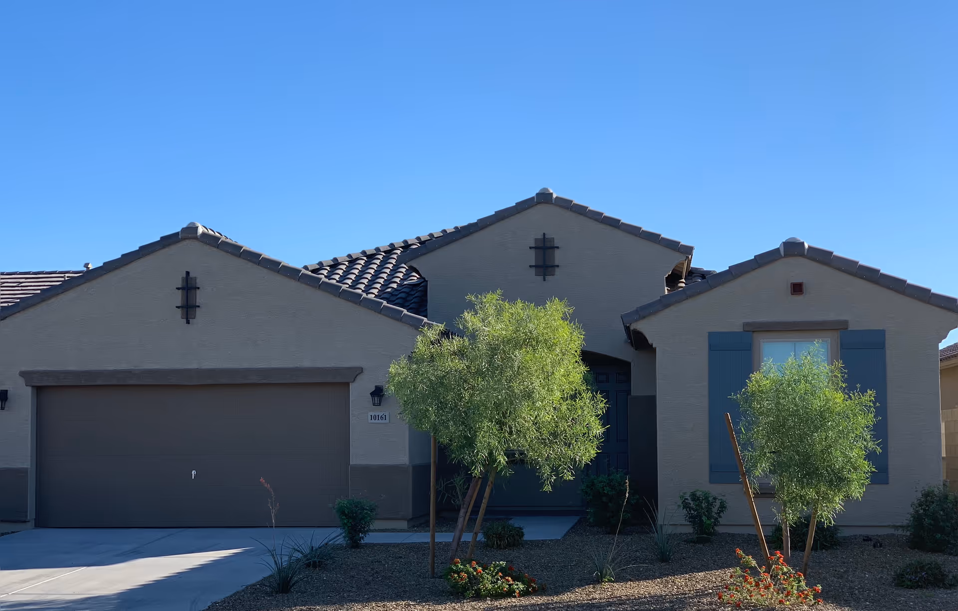 Single-story stucco house front with a two-car garage, tiled roof, and small trees with desert landscaping under a clear blue sky.
