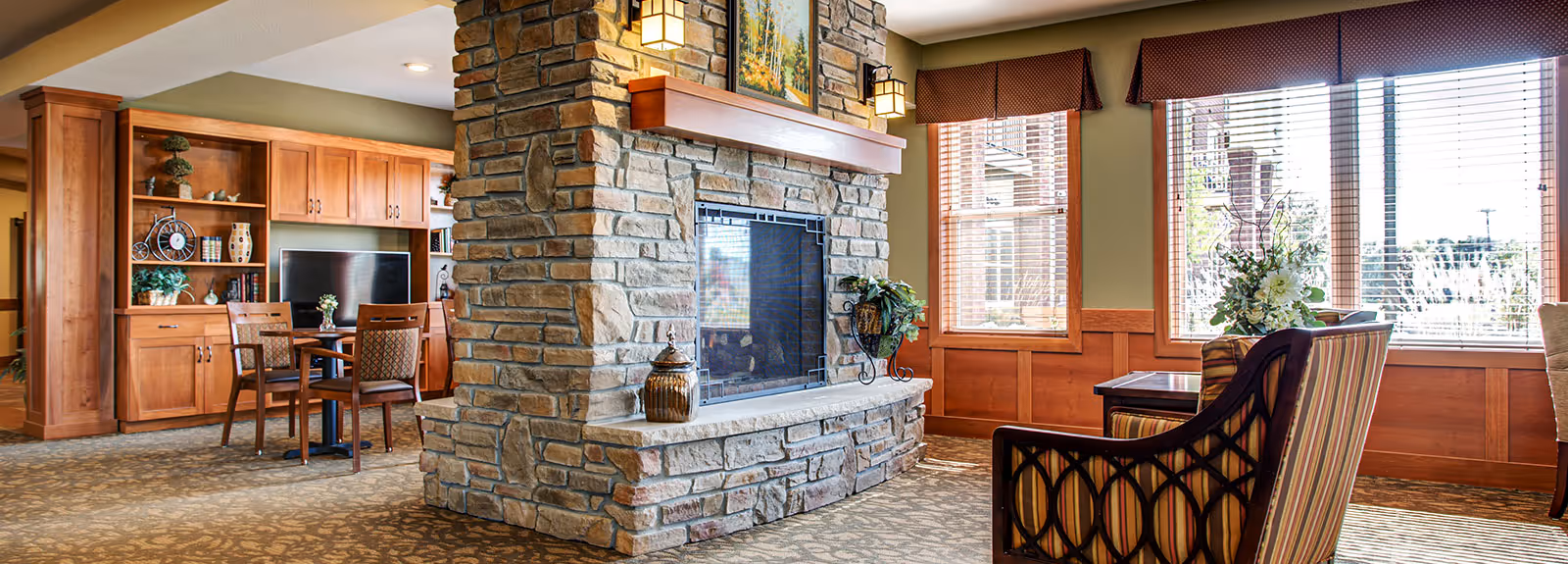 Sunlit senior living common room with a central stone fireplace, seating, wood cabinetry and large windows.