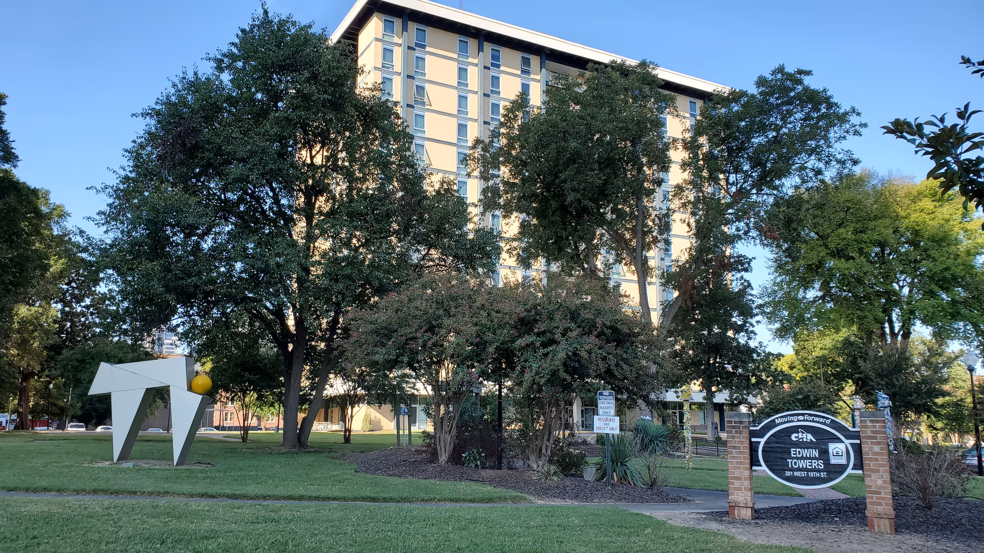 Tall beige senior living building framed by trees, with a modern sculpture and Edwin Towers sign.