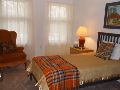 Cozy bedroom in a senior facility with a bed, plaid throw, nightstand, lamp, and armchair.