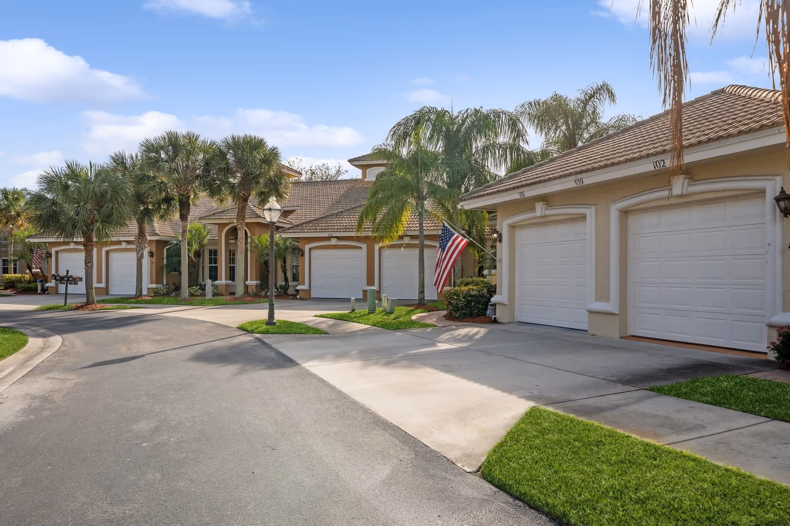 Exterior view of a residential community with beige stucco buildings featuring white garage doors, palm trees, well-maintained lawns, and an American flag displayed near one of the garages under a clear blue sky.