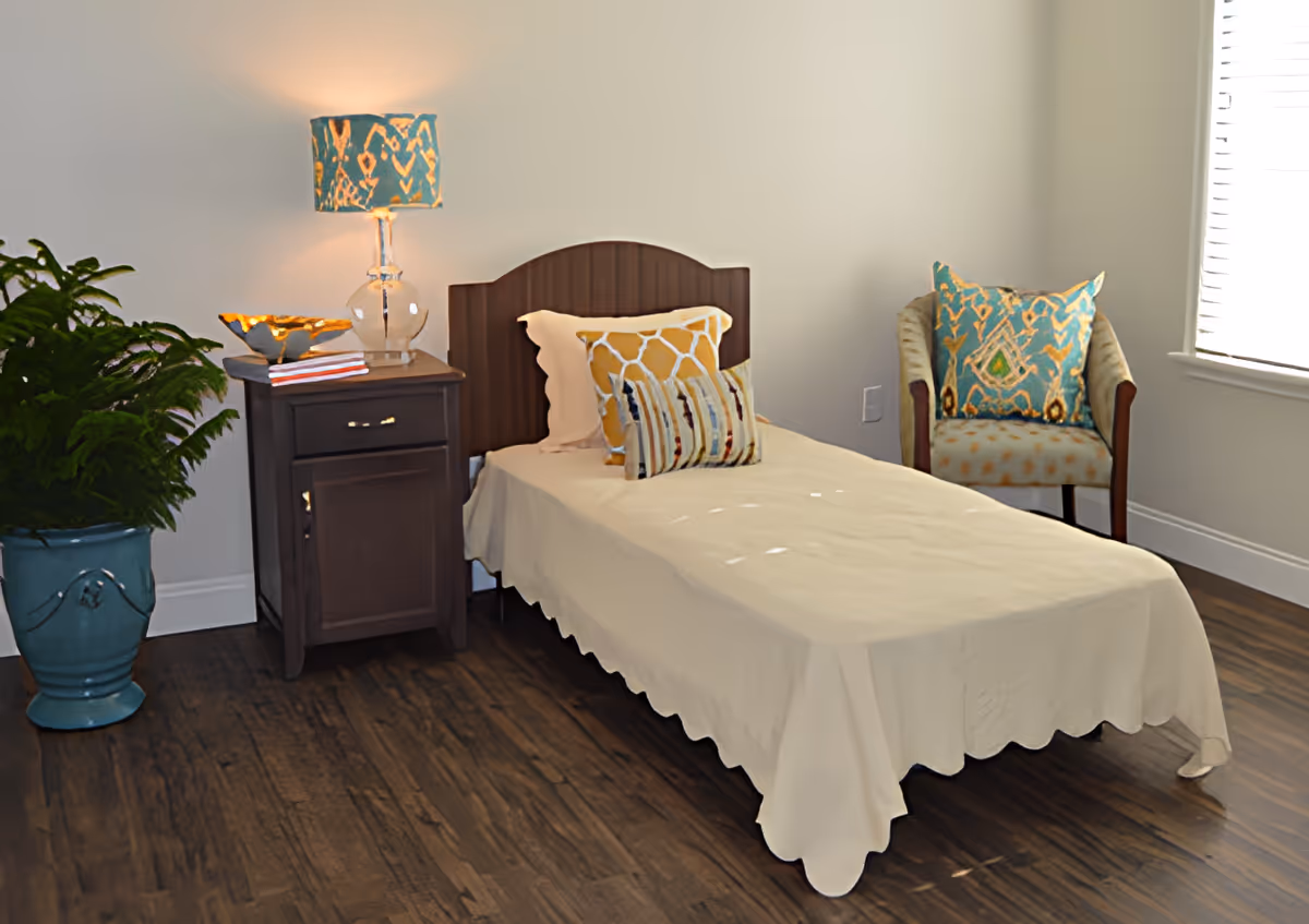 Single bed with a nightstand and lamp, decorative pillows, an upholstered chair, and a potted plant in a bright, simply furnished bedroom.