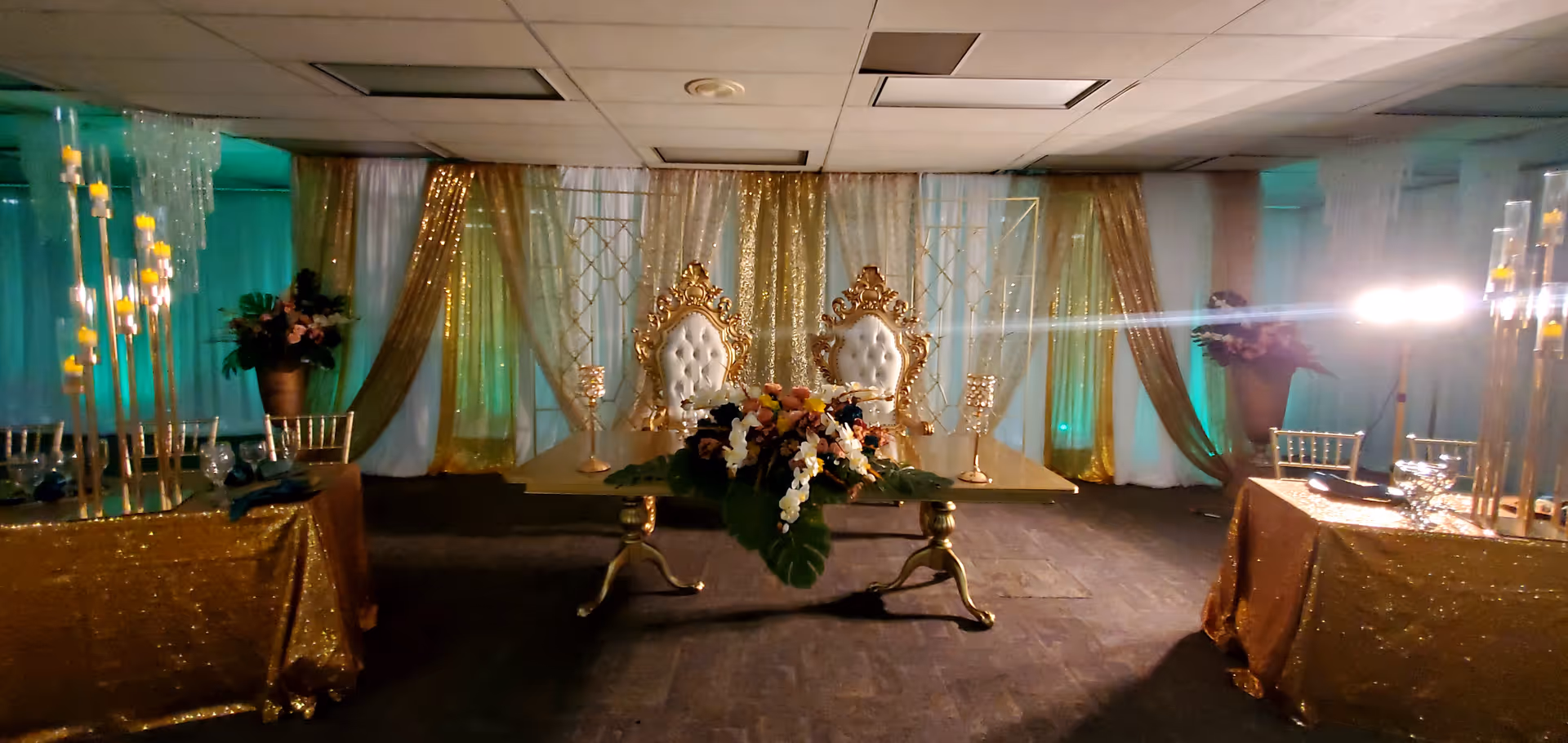 Indoor event hall with gold-draped backdrop, ornate white chairs on a raised platform, and banquet tables.