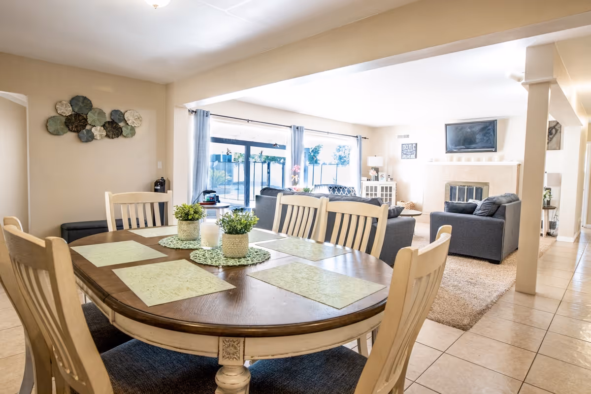 Bright and spacious open-plan dining and living area in a senior living facility. The dining area features a wooden table with six chairs, green placemats, and small potted plants as centerpieces. The living room has two gray sofas, a fireplace with a mounted TV above it, and large windows with sheer curtains allowing natural light to fill the space.