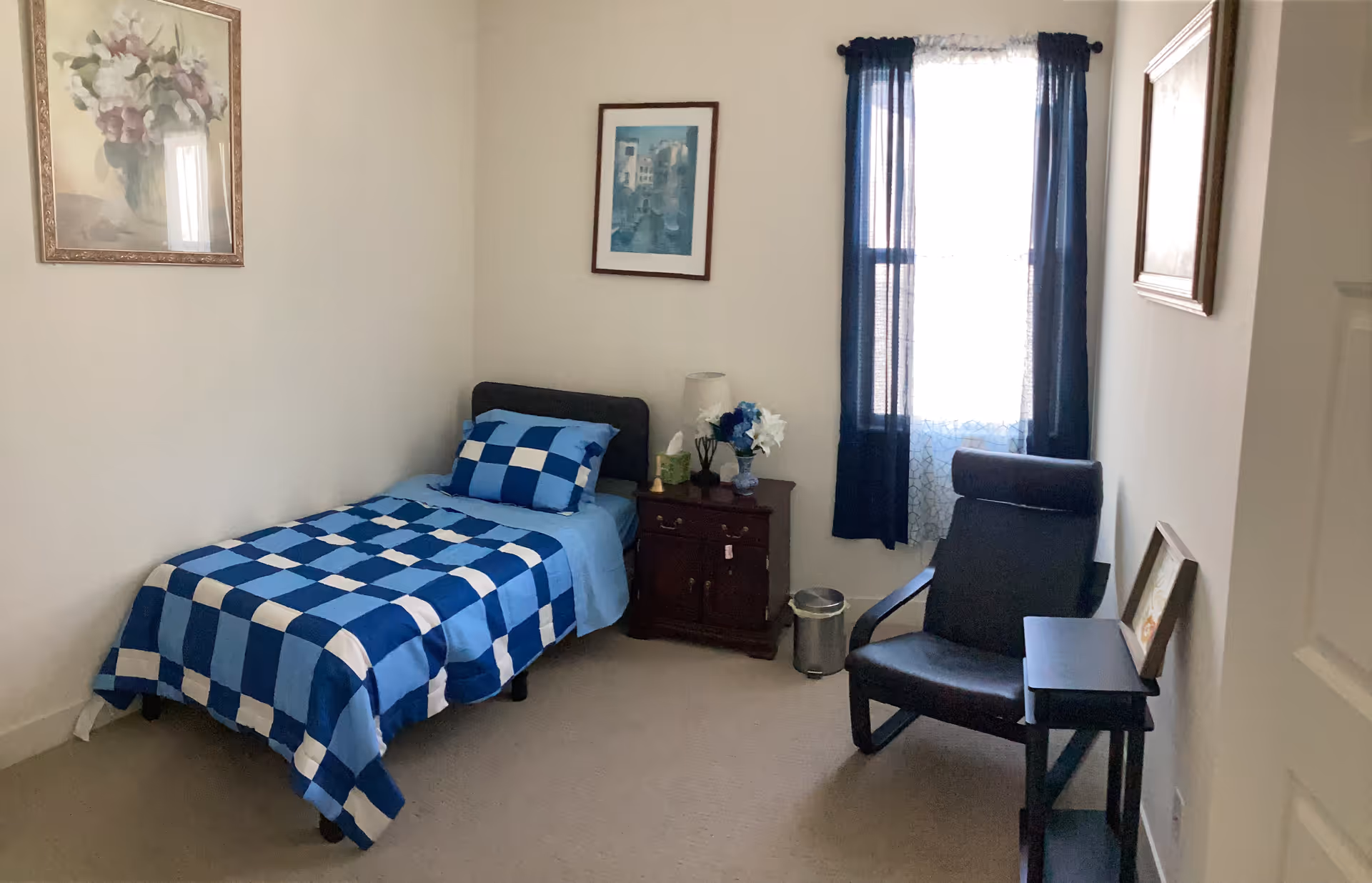 A small bedroom with a single bed, blue checked bedding, a nightstand, and a window.