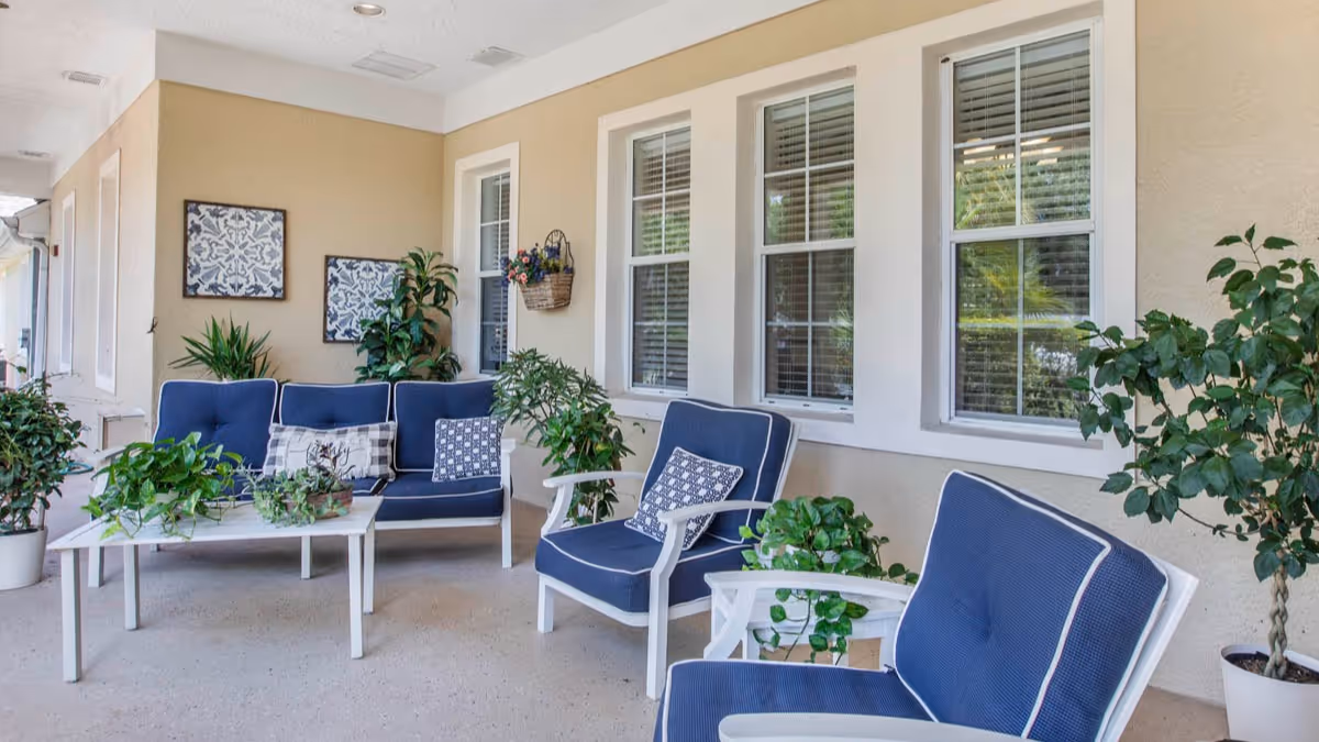 A covered outdoor patio area with white-framed blue cushioned chairs and a matching loveseat around a white coffee table. Several potted green plants are placed on the table and around the seating area. The beige wall behind features three windows with white trim and two decorative framed wall hangings, along with a hanging basket with flowers.