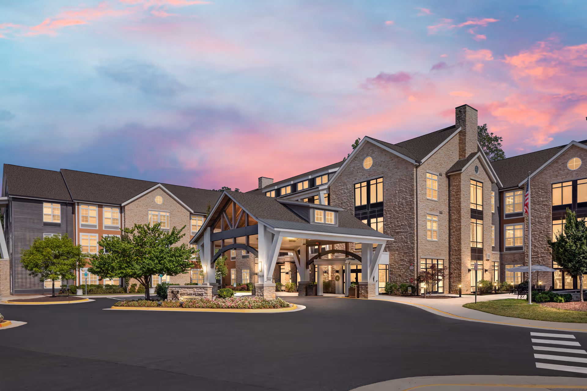 Exterior view of Heritage Village Assisted Living and Memory Care building at dusk, featuring a large covered entrance with wooden beams, well-lit windows, landscaped greenery, and a colorful pink and purple sky in the background.