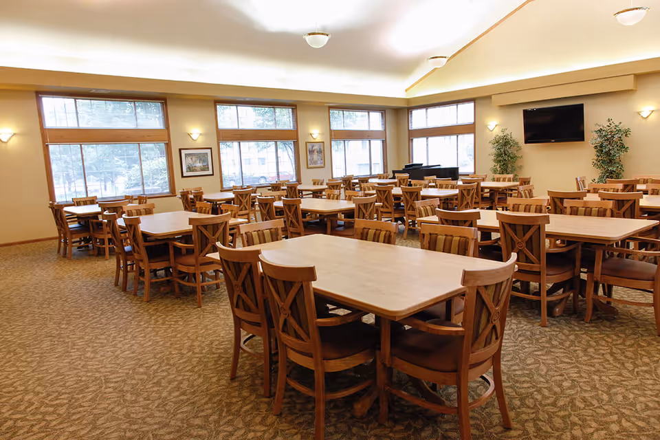 A spacious dining room with multiple wooden tables and chairs arranged neatly. Large windows allow natural light to fill the room. The walls are decorated with framed pictures and there are two potted plants near a wall-mounted television. The carpet has a patterned design and the ceiling has recessed lighting.