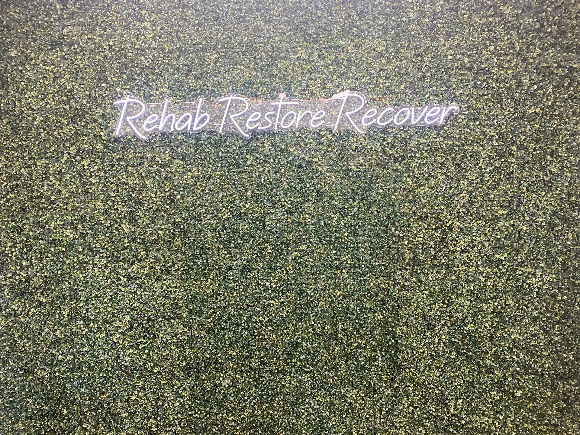 Neon sign reading Rehab Restore Recover on a green leafy wall exterior