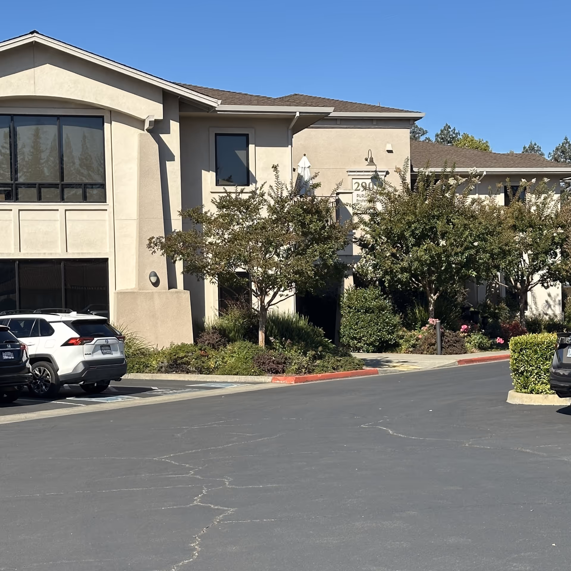 Beige senior care facility exterior with parked cars, trees, and landscaped entrance.