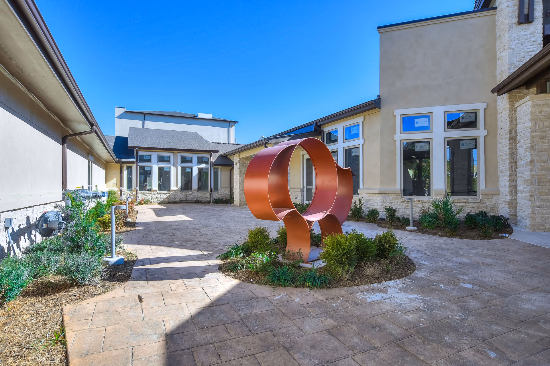 Sunlit courtyard of a senior living facility featuring a large circular metal sculpture, paved walkways, landscaping, and surrounding building windows.