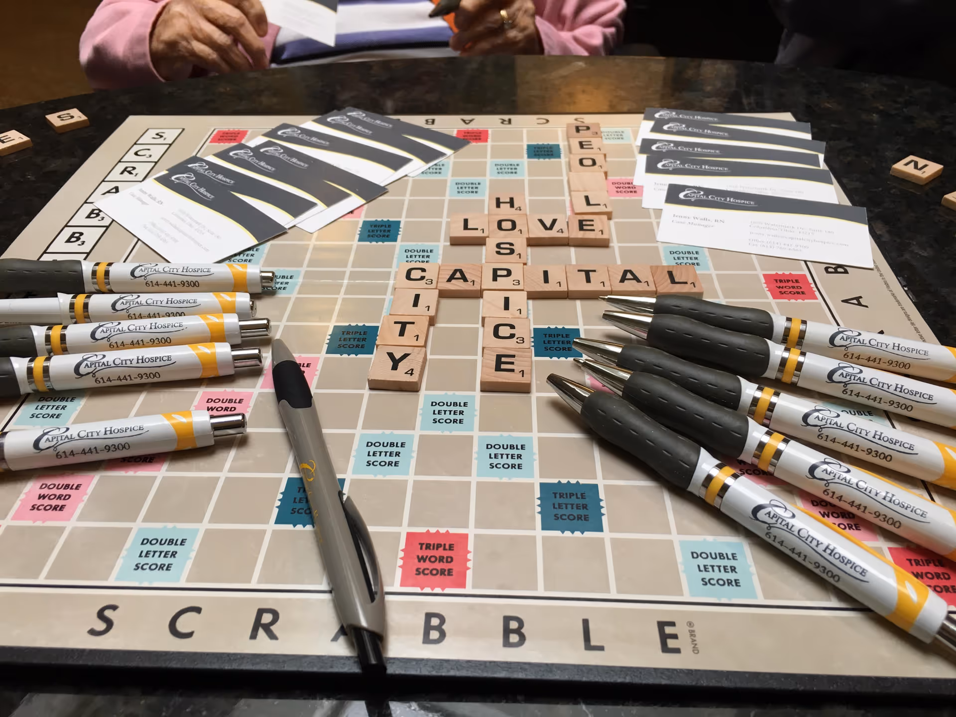 Scrabble board on a table with Capital City Hospice pens and cards visible.