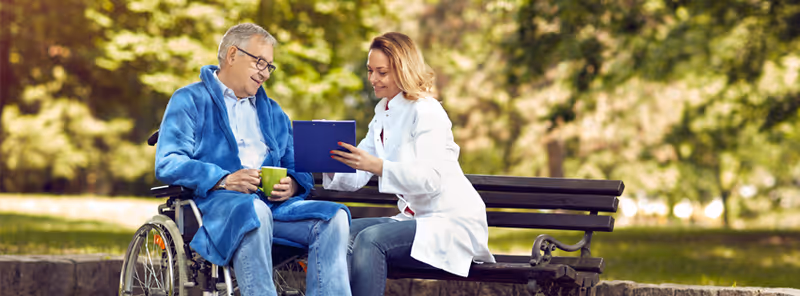 A clinician in a white coat consults with an elderly man in a wheelchair outdoors.