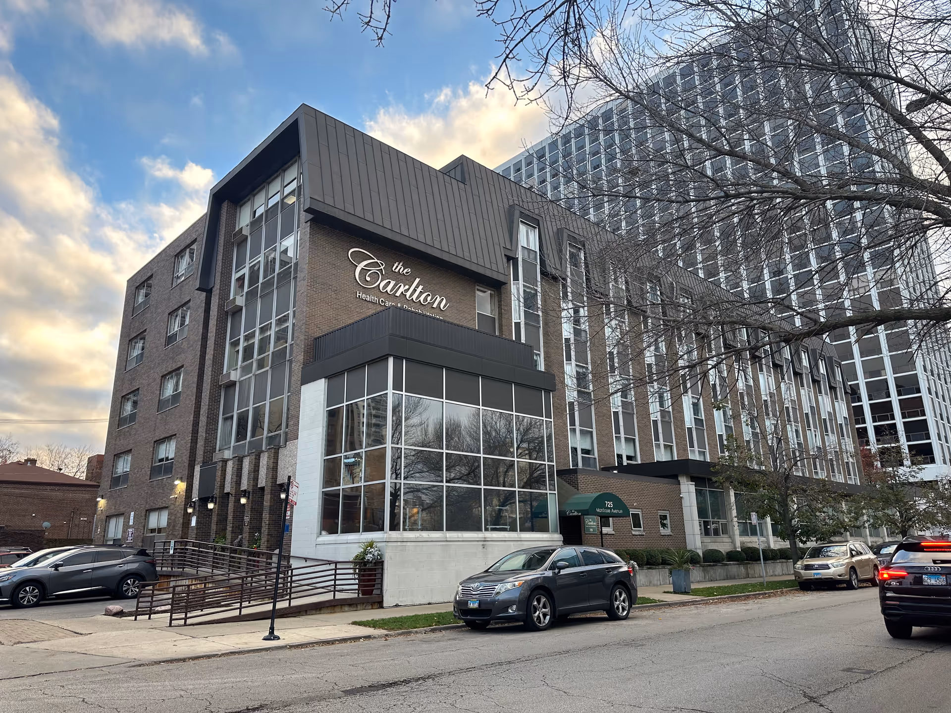 Exterior street view of the Carlton senior living building with a glass-front entrance, brick facade, parked cars, and leafless trees.