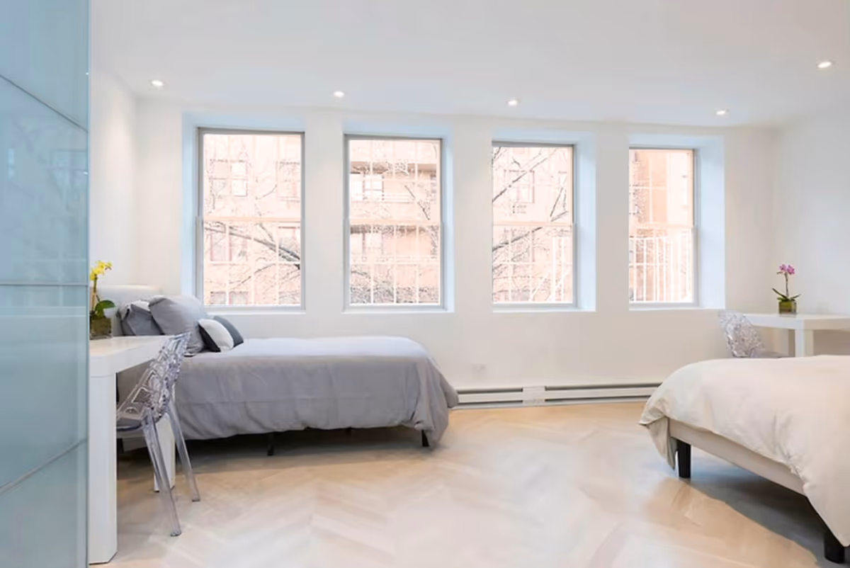 Bright shared bedroom with two beds, desks, chairs, and large windows in a modern facility.