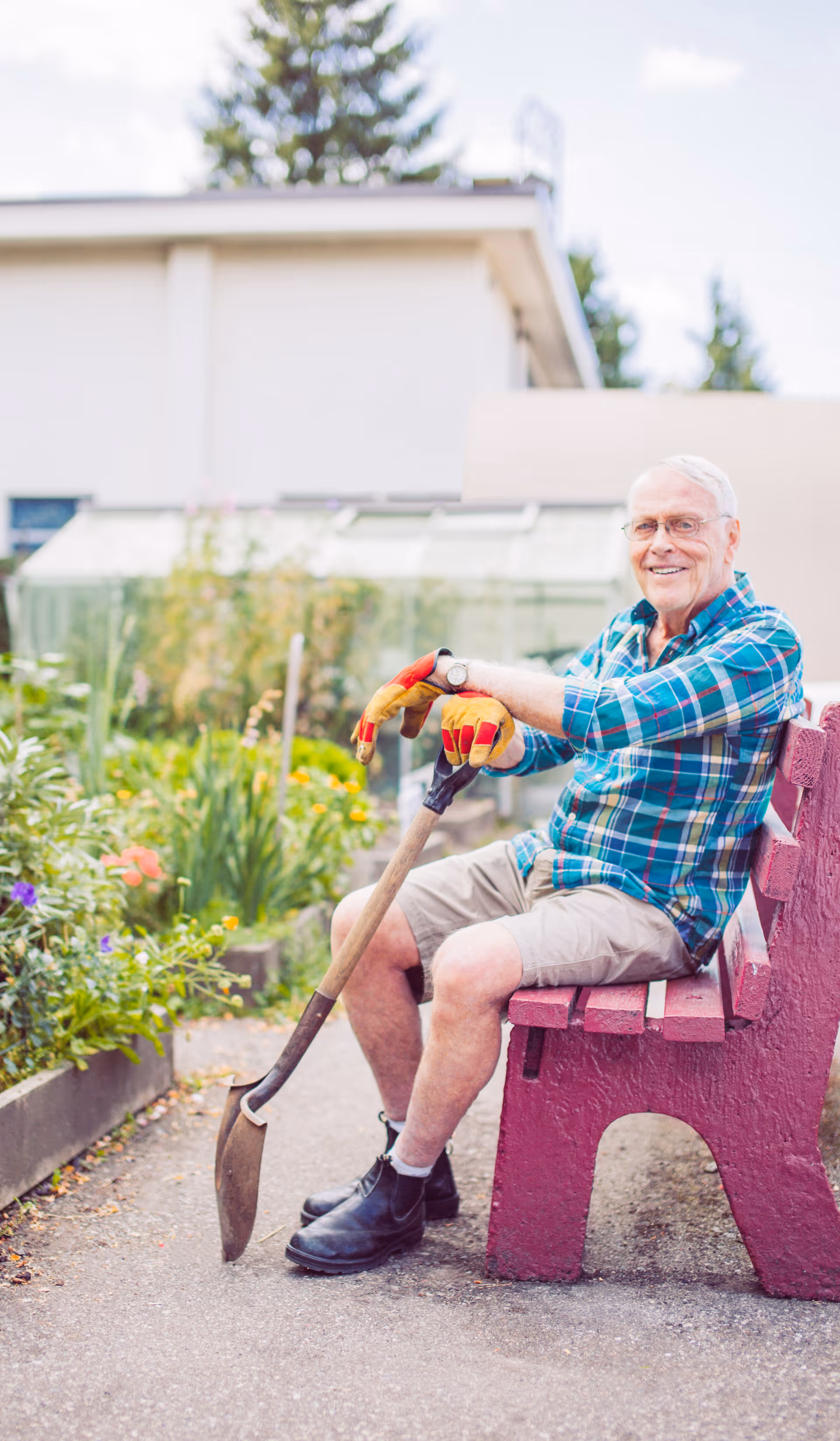 An elderly man sits on a pink bench in a garden, holding a shovel outdoors.