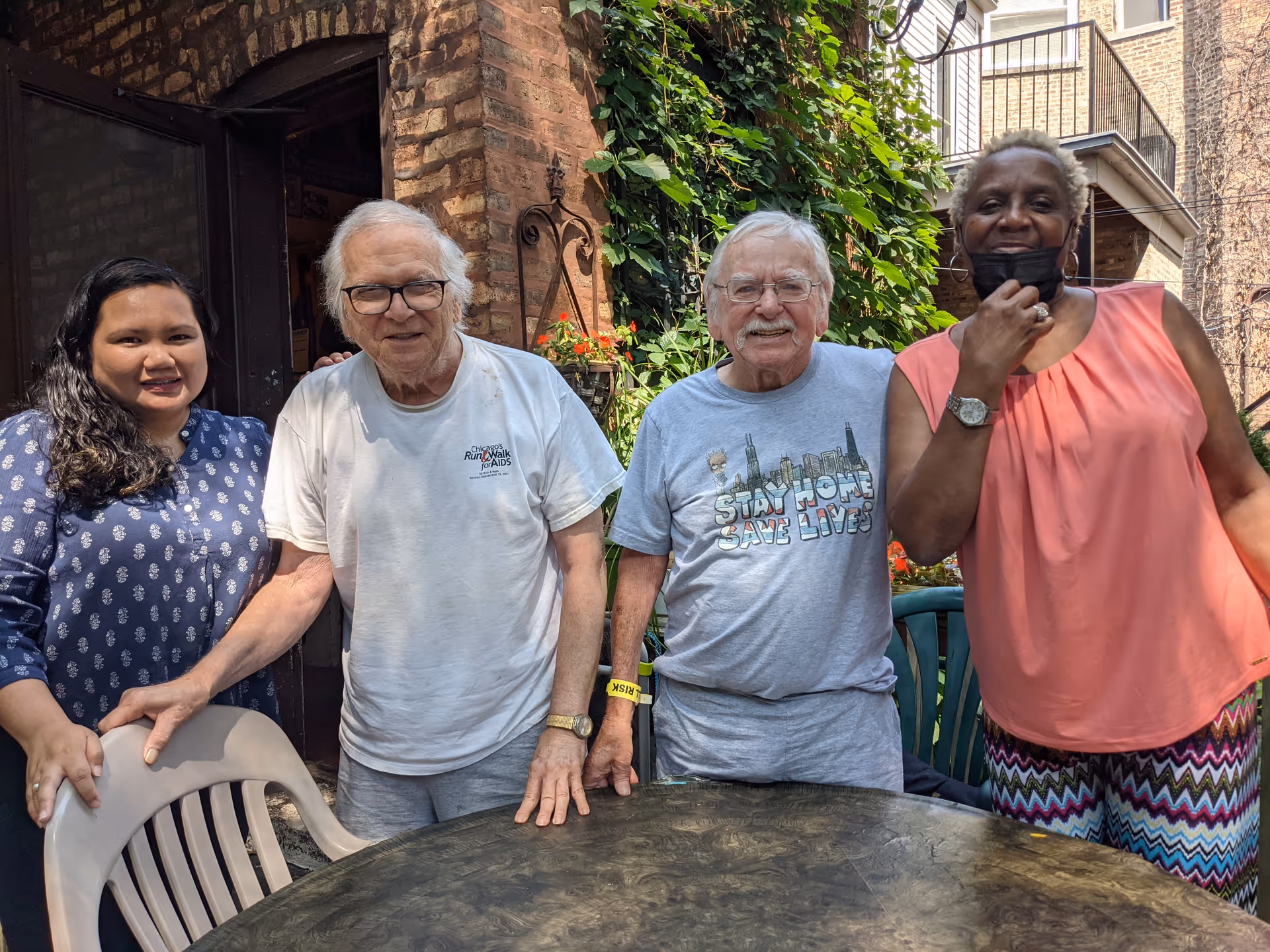 Four adults posing outdoors by a table and chairs near ivy-covered brick wall