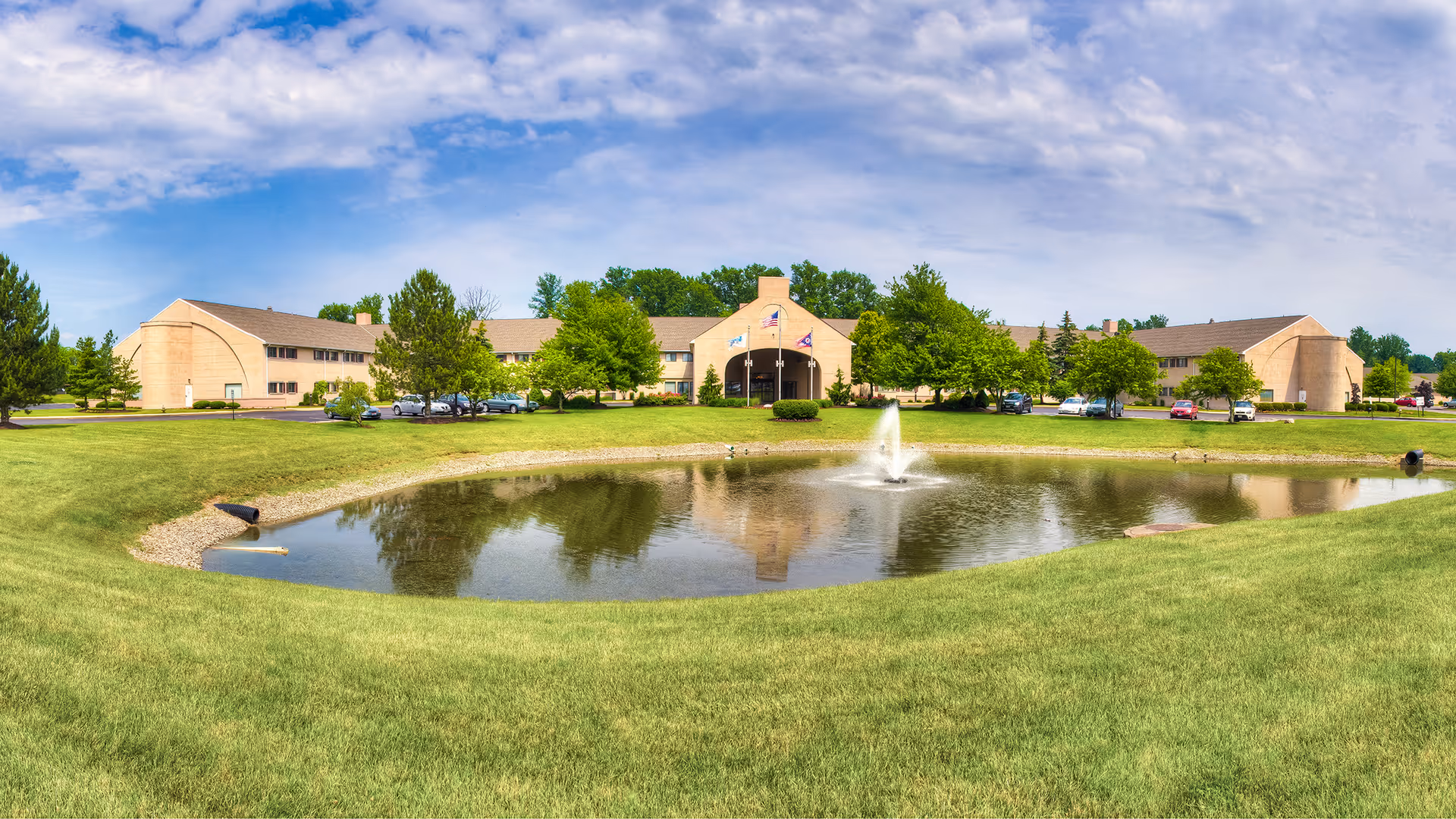 Senior living campus with a pond, fountain, greenery, and a brick front building.