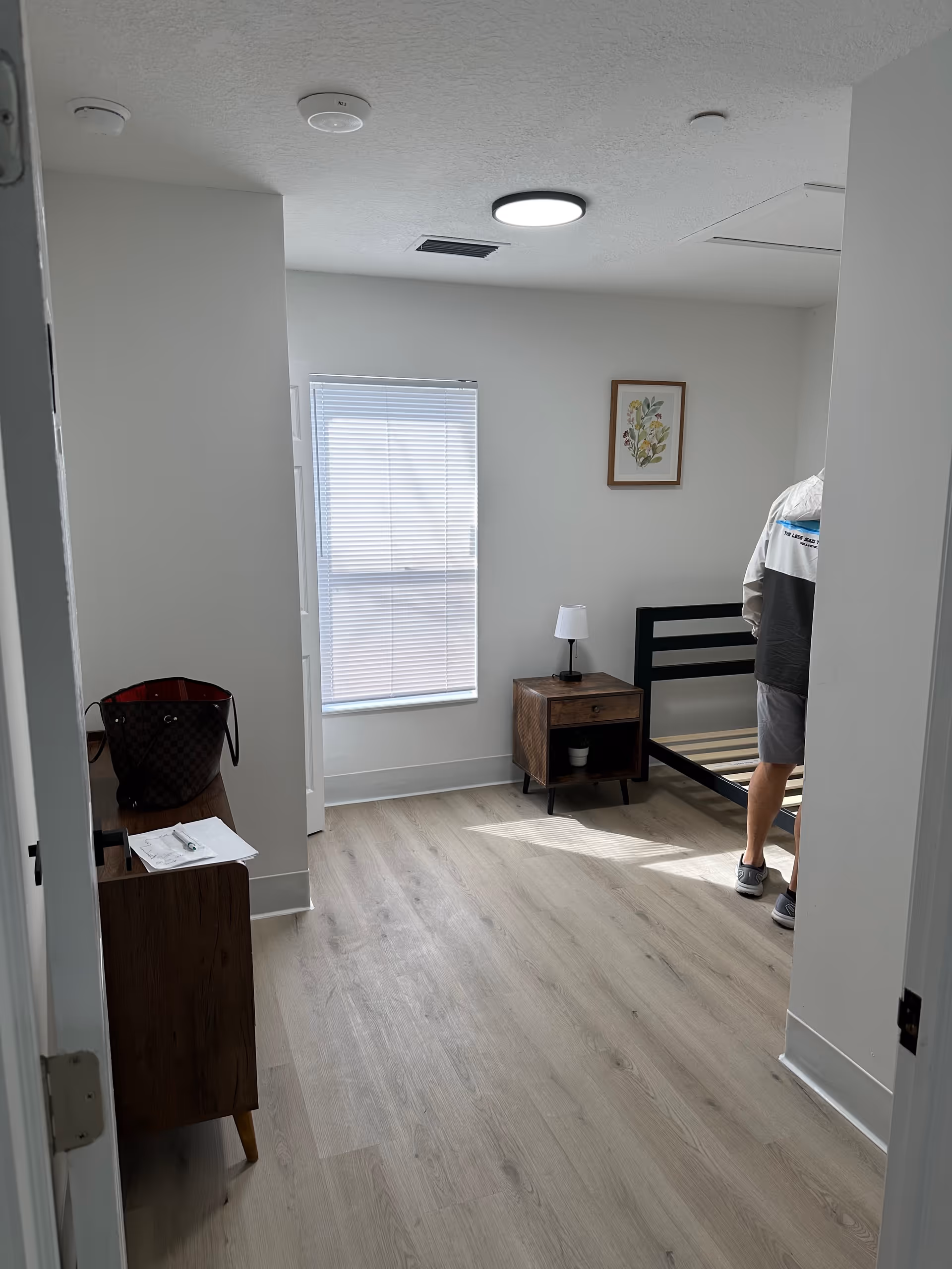 Senior living bedroom with a bed frame, nightstand, lamp, and window blinds.
