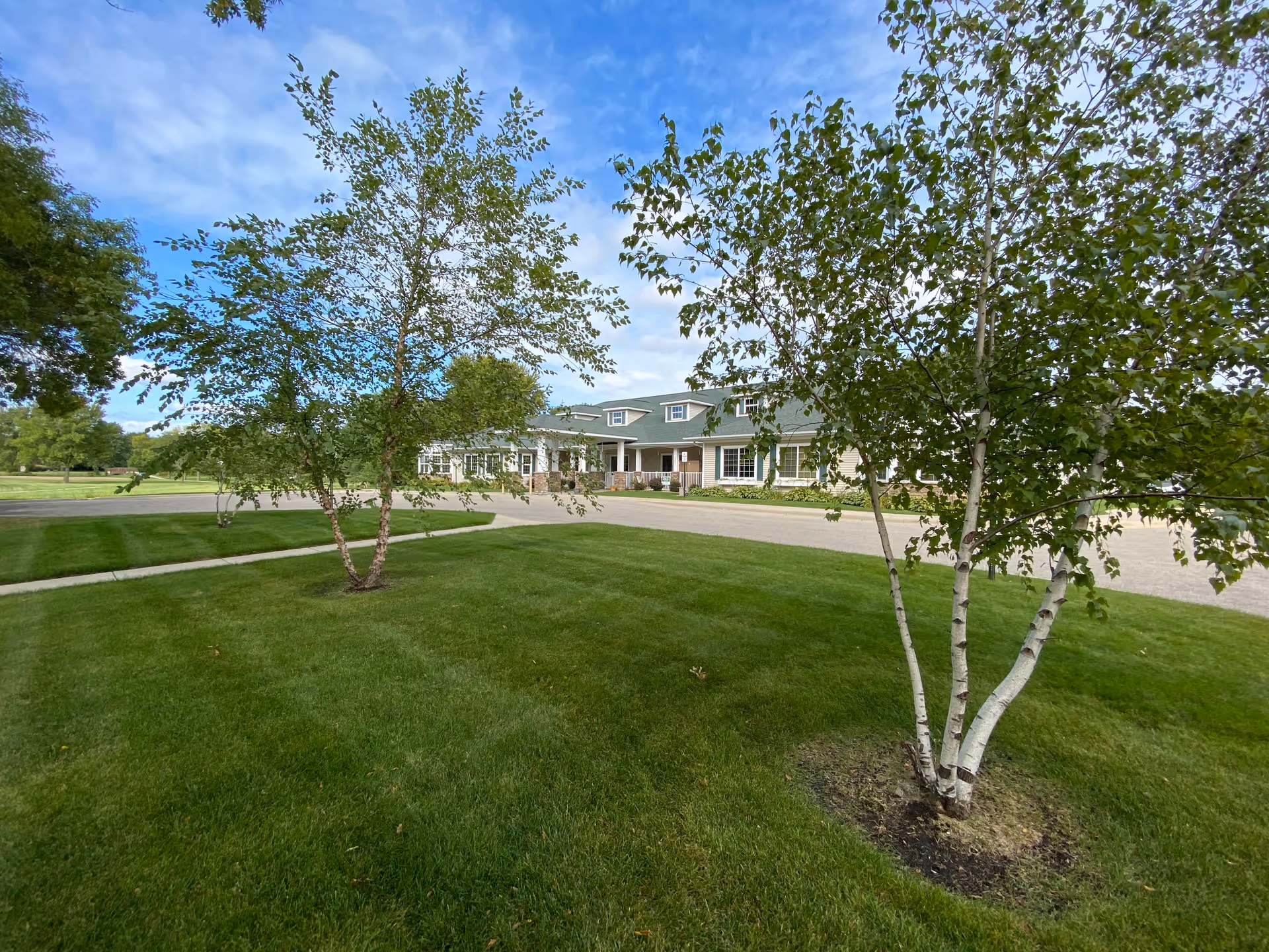 Green lawn with birch trees in the foreground and a single-story senior living building under a blue sky in the background.