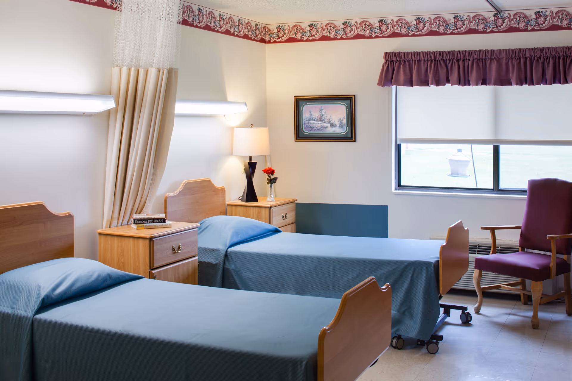 A room with two single beds covered with blue bedspreads, a wooden nightstand with a lamp and a vase with a red flower, a framed picture on the wall, a window with a purple valance and a purple armchair next to it.