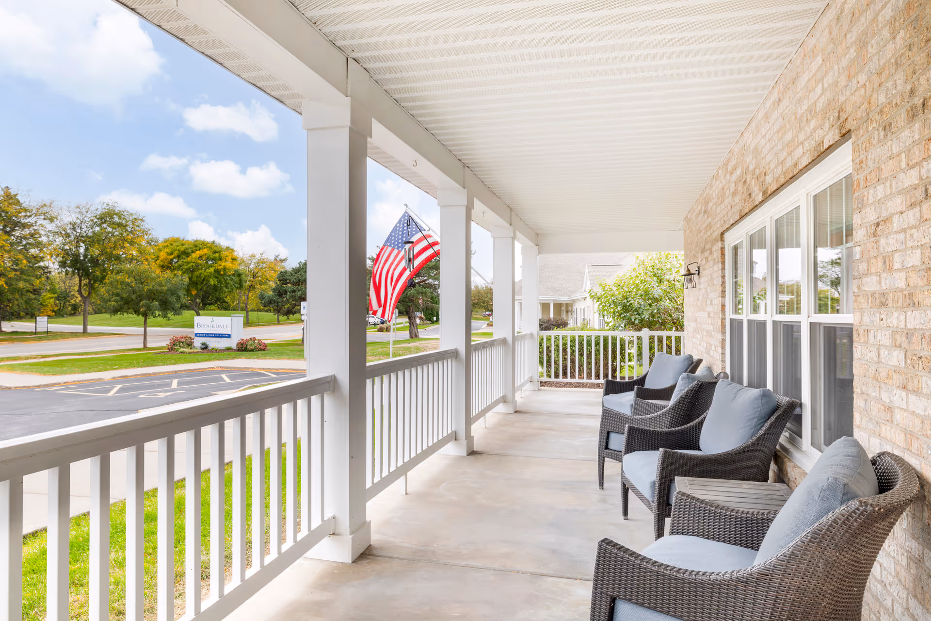 Exterior covered porch of Noelitz Home with wicker chairs, railing, and an American flag.