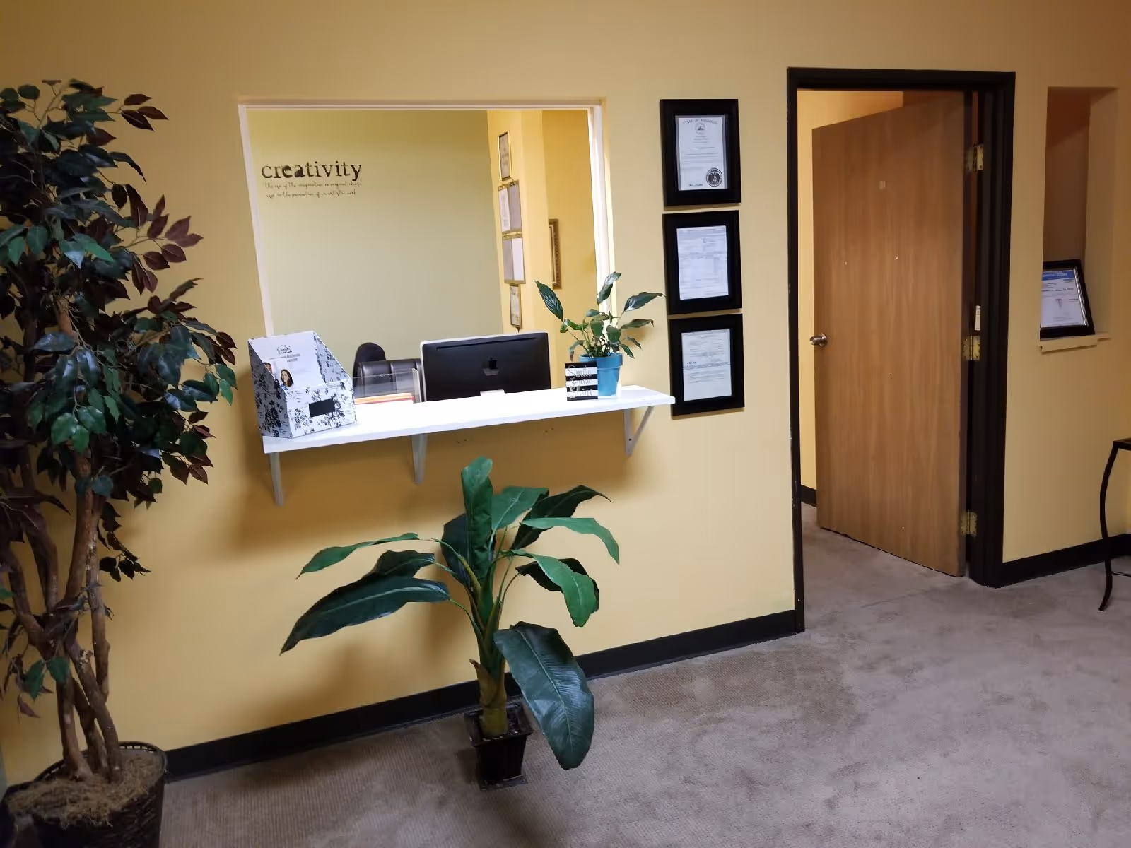 Reception desk with computer, certificates, and potted plants in a yellow-walled lobby.