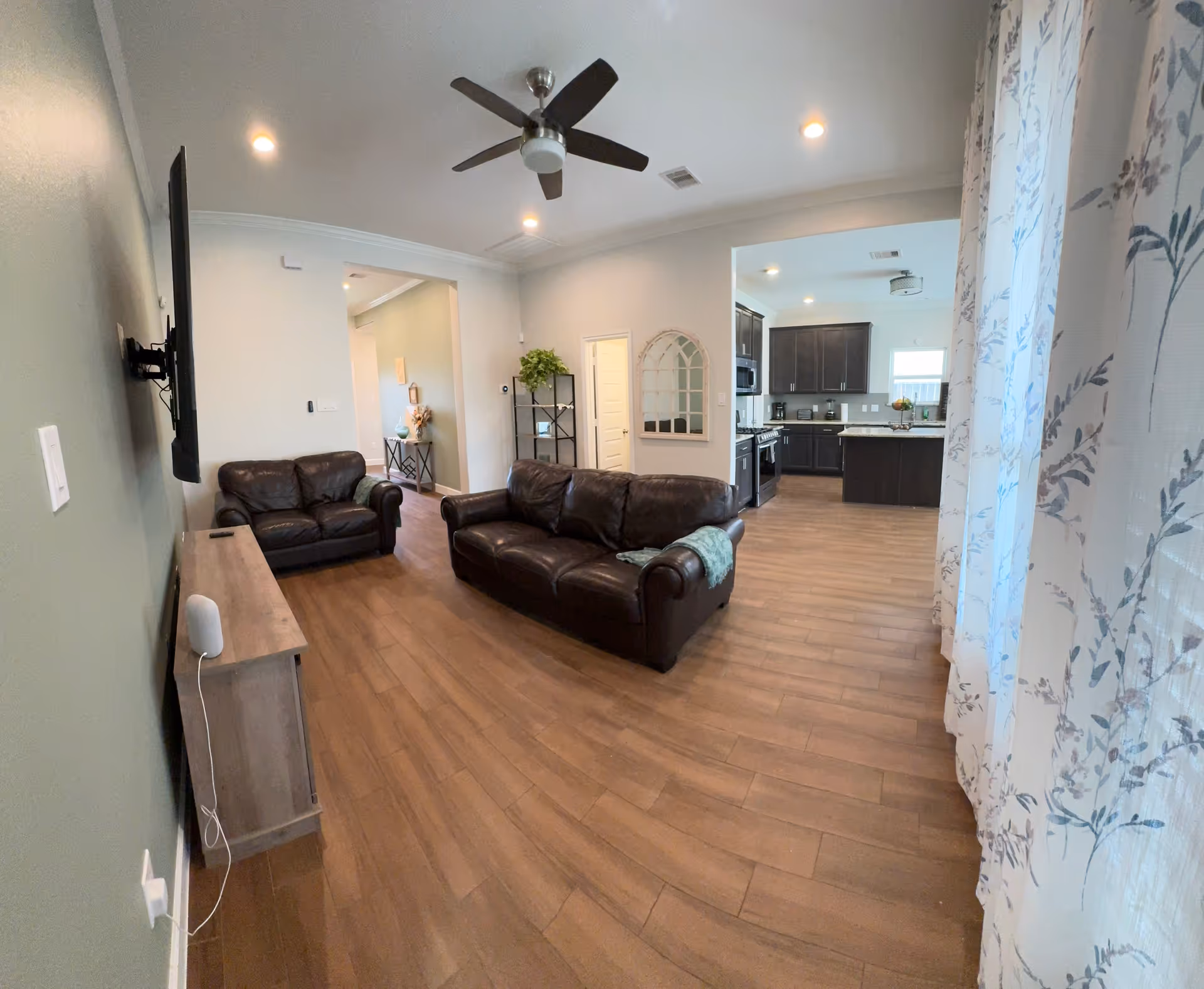 Spacious living room with leather sofas, a wall-mounted TV, ceiling fan, and open kitchen.
