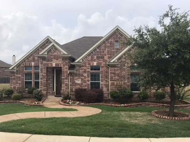 Front view of a red-brick single-story suburban house with gabled roof, a curved walkway, manicured lawn and landscaping.