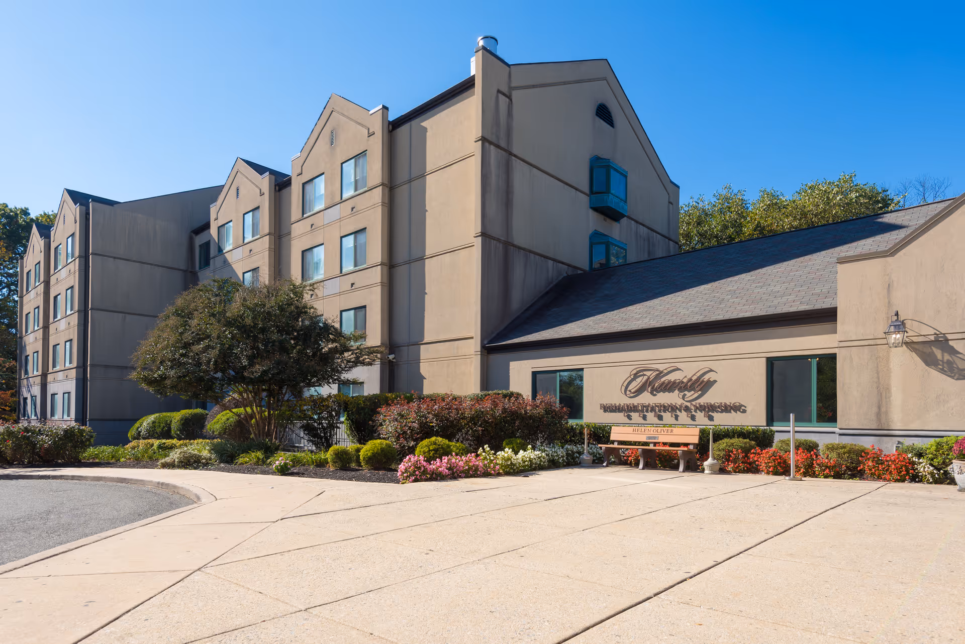 Exterior view of a multi-story nursing center with landscaping and a bench in front.