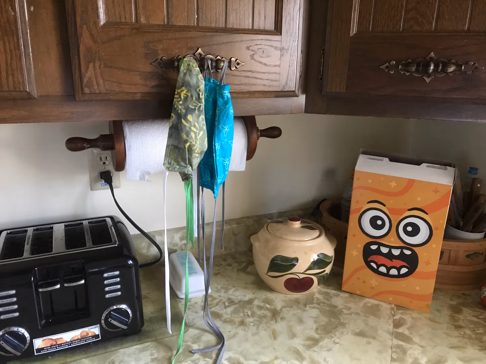 A kitchen countertop with a black toaster, a paper towel holder mounted under wooden cabinets, three hanging face masks, a ceramic container with a lid decorated with leaves and a cherry, and a colorful box with a cartoon face on it.