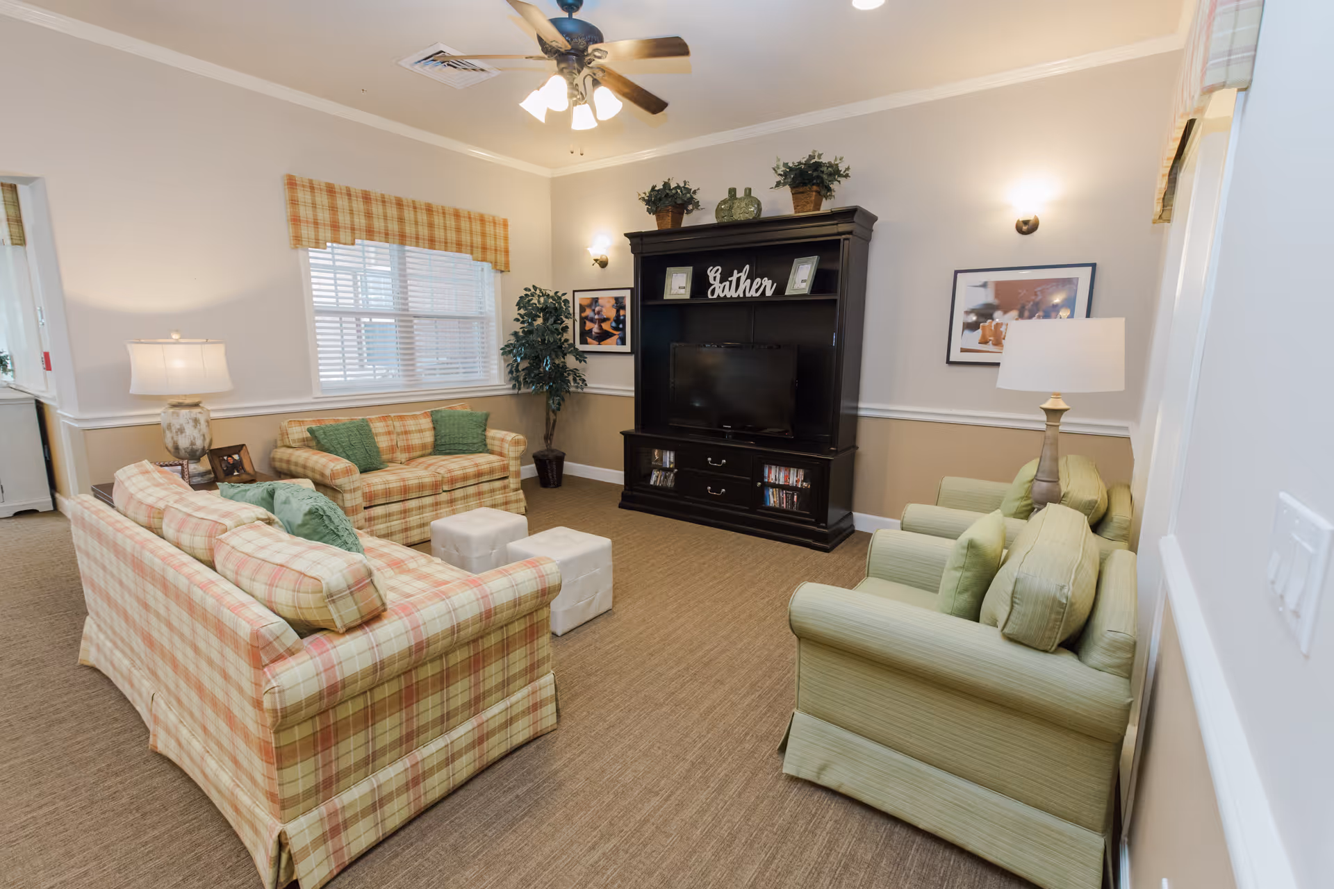 A cozy living room with plaid sofas and green armchairs arranged around a dark wooden entertainment center with a TV. The room has beige walls with white trim, a window with plaid valance, two table lamps, a ceiling fan with lights, framed pictures on the walls, and decorative plants on top of the entertainment center and in the corner.
