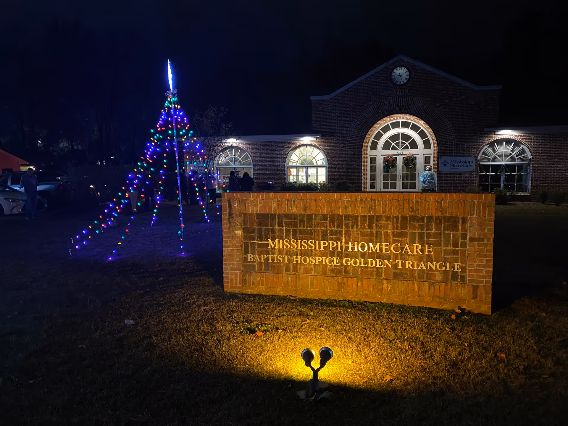 Night exterior of a brick hospice building with a lit Christmas tree and sign
