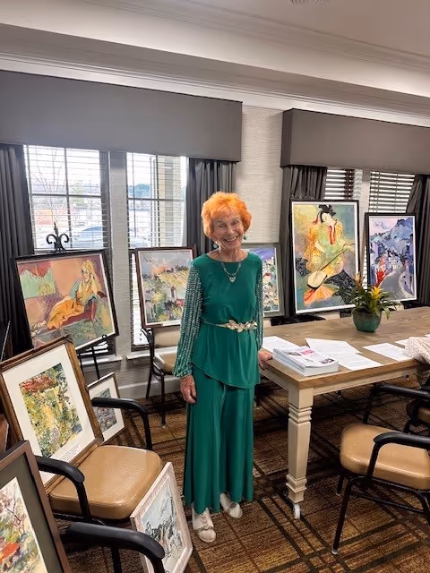 An elderly woman with orange hair wearing a green dress stands in a room with several framed paintings displayed on chairs and easels around her. The room has large windows with gray valances and a wooden table with papers and a potted plant on it.