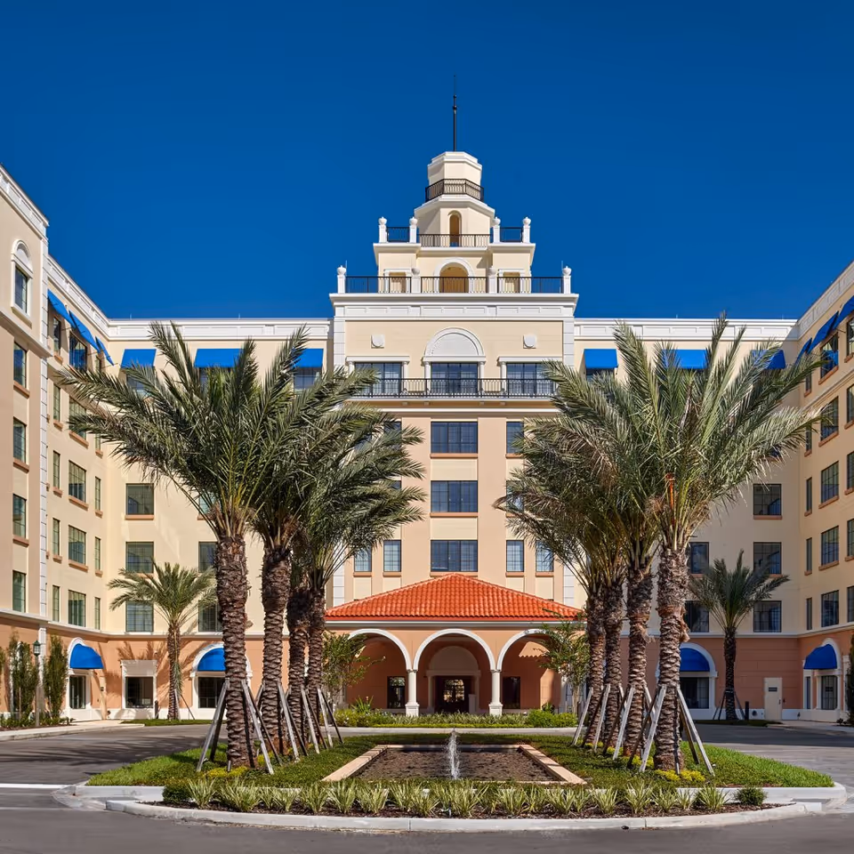 Front exterior view of Windsor at Celebration facility with a symmetrical building featuring multiple floors, palm trees lining a driveway, a small water fountain in the center, and a clear blue sky.