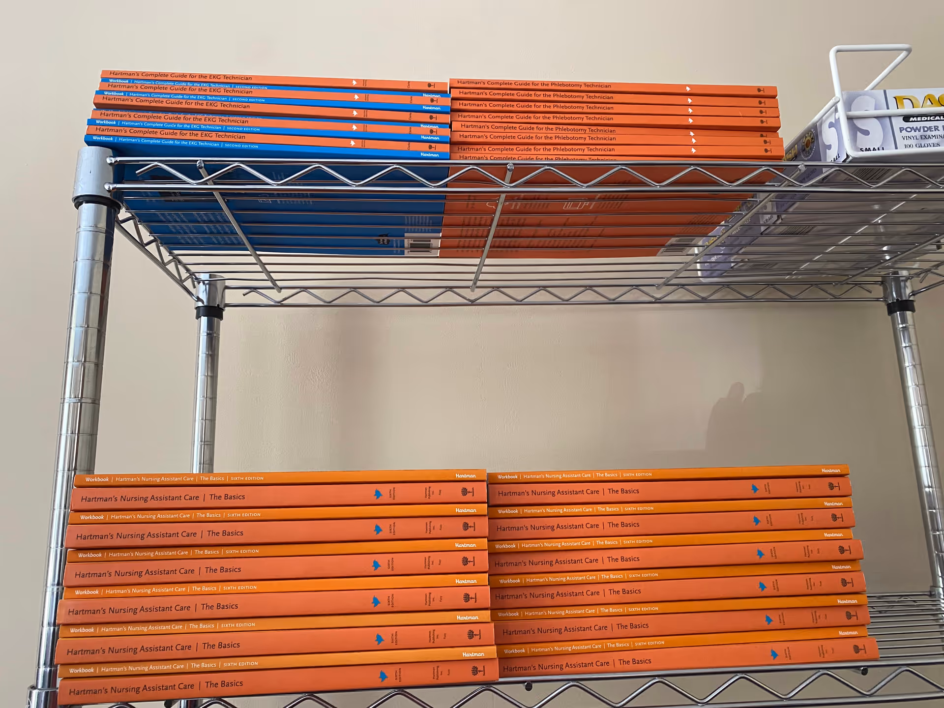 Orange nursing assistant care textbooks stacked on metal wire shelves in a neutral interior.